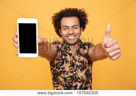 Close up portrait of a happy young afro american man showing blank screen mobile phone and giving thumbs up isolated over yellow background