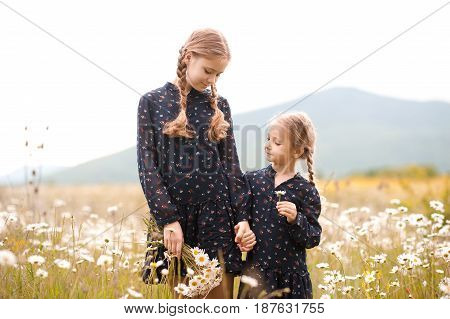 Smiling teen girl 12-14 year old with kid girl 4-5 year old walking in chamomile meadow. Two sisters in similar dresses posing outdoors. Childhood. Summer time.