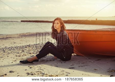 A girl sideways sitting near a red boat on the beach by the sea with beautiful sunset on the background. Woman in jeans pullover and jacket on the sand