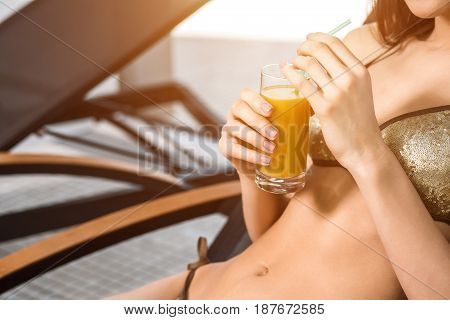 Young attractive slim girl in bikini relaxing on deck chair in wellness spa hotel resort. Woman holding a glass of orange juice. sun flare