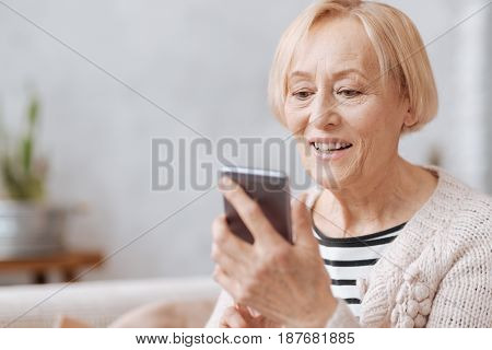 Keeping up with time. Smiling friendly aged lady holding her smartphone in her hands and typing something while spending time at home