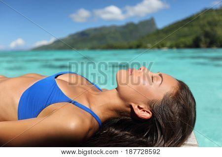 Bikini woman relaxing sunbathing on luxury resort on overwater deck. Sexy Asian model lying down sun tanning on turquoise ocean Tahiti water.