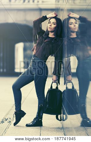 Happy young woman standing at the wall in city street. Stylish fashion model in leather fringe suede jacket and blue jeans