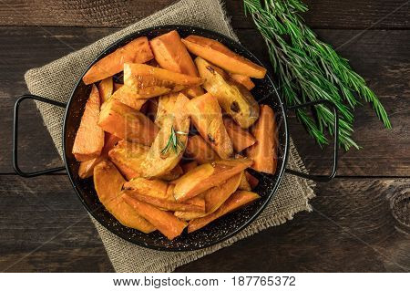 An overhead photo of roasted sweet potatoes in a pan, shot from above on dark rustic wooden textures with rosemary branches, with a place for text