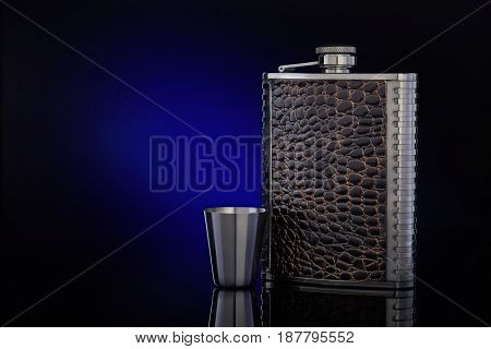 Hip flask from stainless steel with brown leather  for whisky or cognac and steel drinking glass on a dark blue background with copy space.