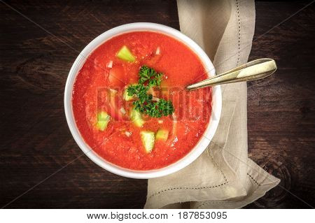A bowl of gazpacho soup with slices of tomato, cucumber, and onion, with parsley leaves, shot from above on a dark rustic texture with a place for text