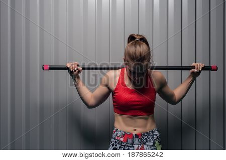 Portrait of young sporty woman wearing red top with bodybar on shoulders. Sports girl posing against grey wall. Fitness, sports and healthy lifestyle.