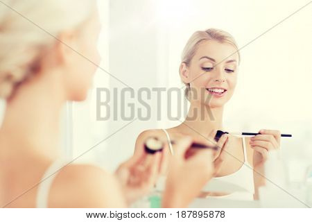 beauty, make up, cosmetics, morning and people concept - young woman applying eyeshade with makeup brush and looking to mirror at home bathroom