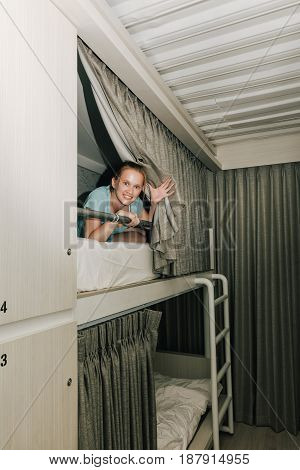 Girl sitting in a stylish hostel bedroom. Young woman sitting on second level bed in hotel room with two-levels beds. Evening grey curtain and wall