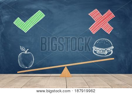 A wooden seesaw on blue chalkboard background where a painted apple overweighs a sandwich. Healthy eating. Staying in shape. Choose your diet.