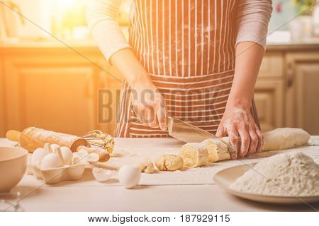 Close-up view of two woman's hands cut knife dough. A woman in a striped apron is cooking in the kitchen
