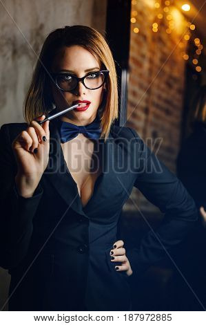 Young attractive girl in a jacket bow tie and glasses. Femme fatale. Evening makeup smokey eye. She lustfully bites the pen.