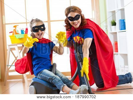 Kid girl and her mother weared Superhero costumes. Cute helper child and woman make cleaning room and have a fun pastime.
