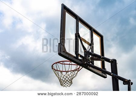 Transparent rectangular fiberglass backboard and red basketball hoop/ rim & white net/ netting. Outdoor urban school basket ball court. Children's community street sports park. Team sport competition.