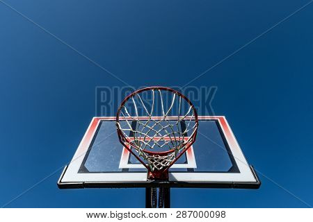 Transparent rectangular fiberglass backboard and red basketball hoop/ rim & white net/ netting. Outdoor urban school basket ball court. Children's community street sports park. Team sport competition.