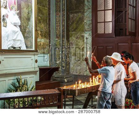 Havana, Cuba - 25 July 2018: Old Man In A Church In Cuba Praying To His Lord Holding Two Lit Candles