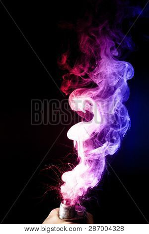 Male Hand Holds Vape. Vape Clouds Splash From Vape Coil On Black Background. Fog Is Dual Color -- Bl