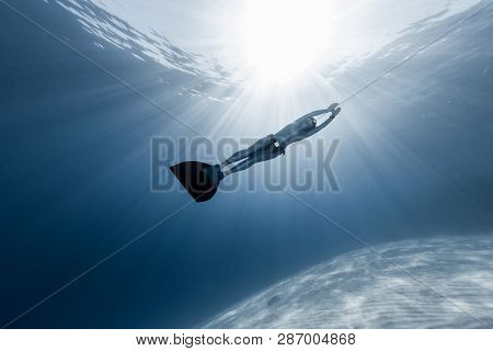 Woman freediver in monofin glides in the depth over the sandy bottom with sunny rays shining through the water