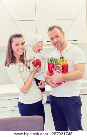 Happy Parents Standing In The Kitchen And Prepares For Cooking With Their Baby Girl Chef. Father Bri