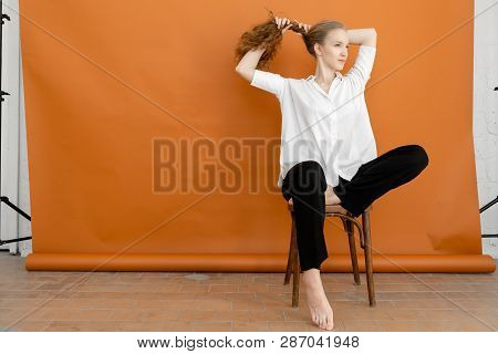 Beautiful Model On Orange Background Sitting On The Chair In White Shirt And Black Trousers