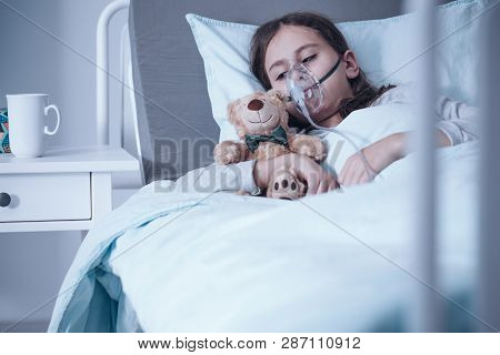 Sad Kid With Cystic Fibrosis Lying In A Hospital Bed With Oxygen Mask And Plush Toy