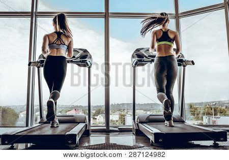 Back View Of Sports People On Running Track. Two Attractive Young Women Are Running In Gym. Treadmil