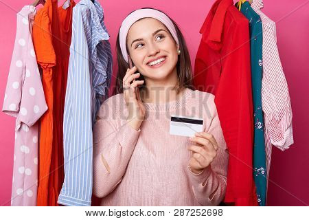 Young Woman With Credit Card Dreams About Shopping While Standing Near Lots Hangers With Colourful C
