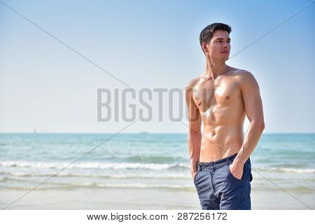 Handsome Young Man With Nude Torso Standing And Looking Sideways Over Beach Background. Copy Space.