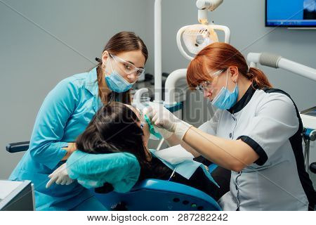 Dentist Woman And Her Assistant In Medical Clothes Treating Patients Teeth In A Stomatological Centr
