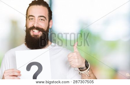 Young man with long hair and bear holding a paper with question mark happy with big smile doing ok sign, thumb up with fingers, excellent sign