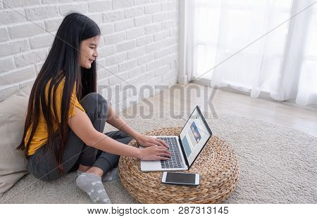 Asian Female Sit With Knees Up On Floor Using Laptop See Social Media Website On Wicker Stand Near W