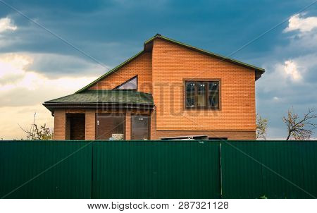 Brick House Under Green Fence With Attic, Windows And Shingles Roof
