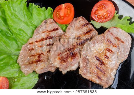 Top View Of The Grilled Sappy Pork Neck Steaks With Lettuce Leaves And Cherry Tomatoes On Black Dish
