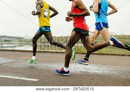 Two African And European Runners Running Marathon In Group Along River Embankment