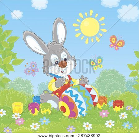Little Grey Bunny Coloring A Big Easter Egg On Green Grass Among Flowers On Its Front Lawn Near A Sm