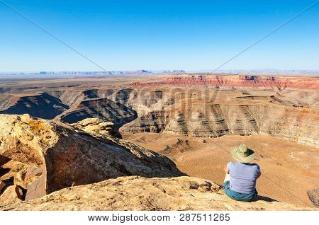 Female Hiker Sitting On The Edge Of Muley Point Overlook With Panoramic View Of The Desert Between U