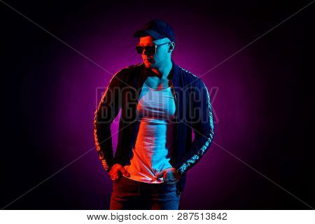 Portrait Of A Young Happy Serious Man In Cap, White Shirt, Bomber And Sunglasses At Studio. High Fas