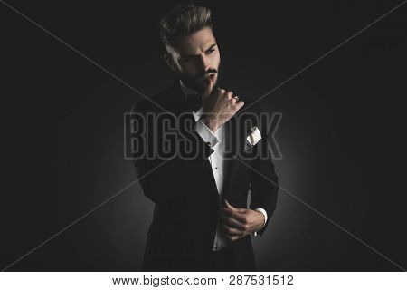 man in tuxedo with bowtie and white hankerchief adjusting his lounge jacket with his thumb at lips and thinking on black background