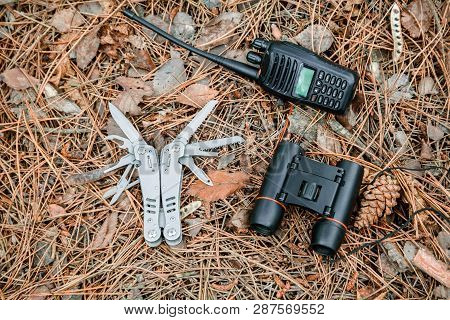 Binoculars, Radio Set And Multitool Knife On The Ground Covered With Pine Needles