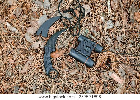 Binoculars And Karambit Knife On The Ground Covered With Pine Needles