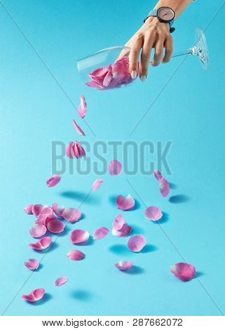 Delicate pink rose petals are pouring from transparent glass which girl's hand holds with wirstwatch on it. Concept production of wine from natural products.