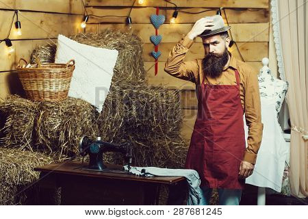 Handsome Man Tailor Or Dressmaker With Beard And Moustache In Apron Puts Hat On Head Near Vintage Se