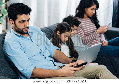 Selective Focus Of Handsome Latin Man Holding Remote Contol Near Hispanic Hamily Using Gadgets