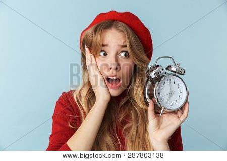 Image of uptight blond woman 20s wearing red beret holding alarm clock isolated over blue background in studio