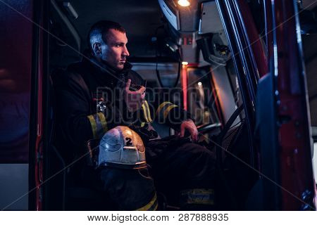 The Fire Brigade Arrived At The Night-time. Serious Fireman Sitting In The Fire Truck And Talking On