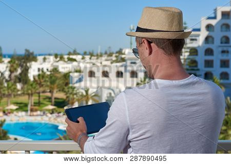 European Man In A White T-shirt And Sun Hat Is Taking Photo Of A Picteresque Hotls Veiw Using Tablet