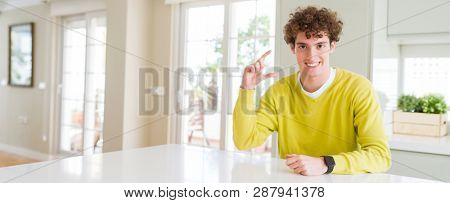 Wide angle shot of young handsome man at home smiling and confident gesturing with hand doing size sign with fingers while looking and the camera. Measure concept.