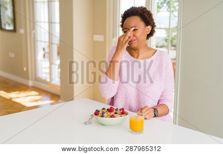 Young african american woman having healthy breakfast in the morning at home smelling something stinky and disgusting, intolerable smell, holding breath with fingers on nose. Bad smells concept.