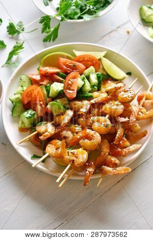 Prawns Skewers And Vegetables Salad From Sliced Cucumber And Tomatoes On White Plate Over Wooden Tab