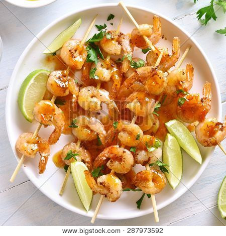 Prawns Skewers With Greens, Spices, Lime And Sauce On White Plate Over Wooden Table. Grilled Shrimp 